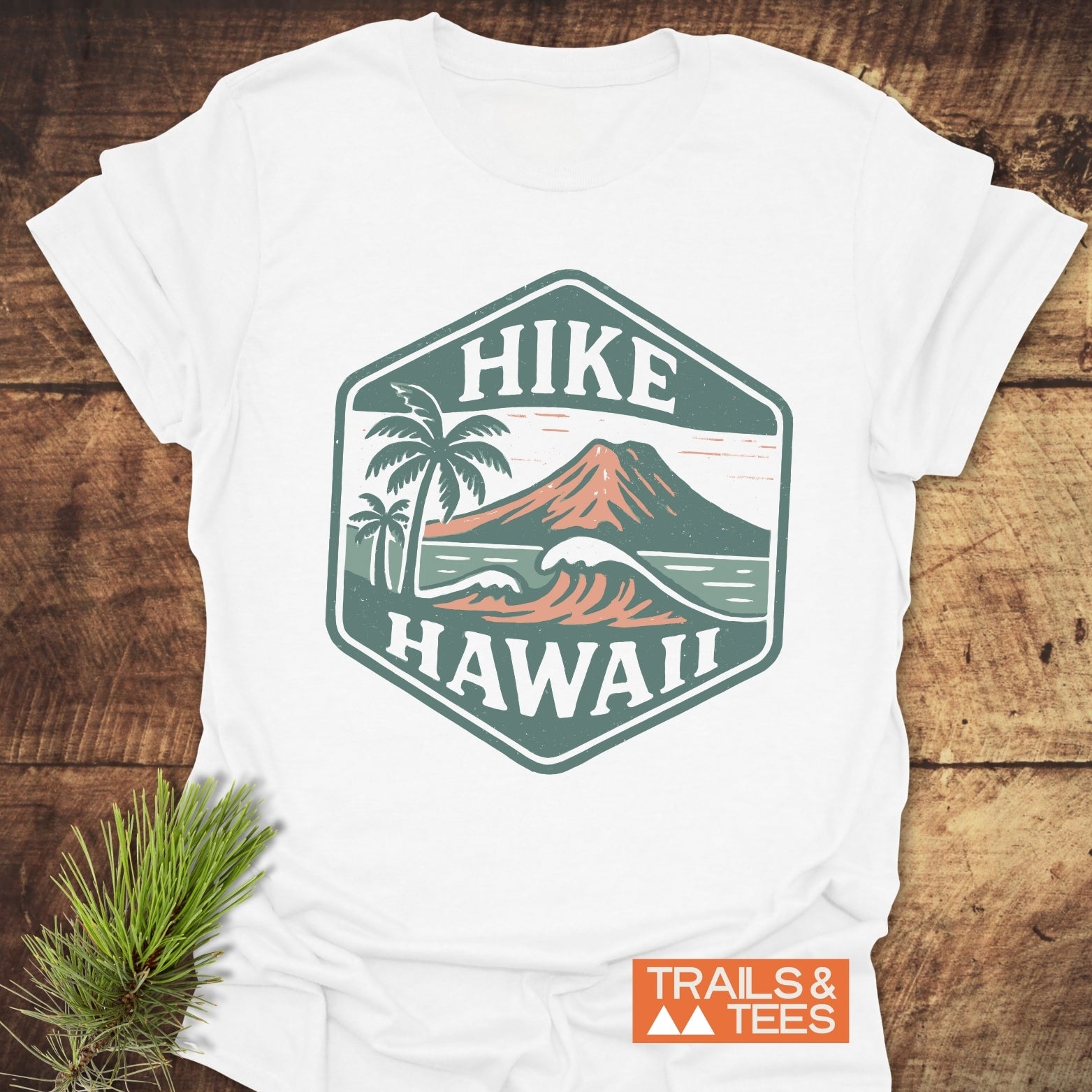Hike Hawaii - Softstyle T-Shirt featuring a retro hexagonal badge with a volcano, ocean waves, and palm trees, laid on wood with a pine sprig. Includes Trails & Tees logo.