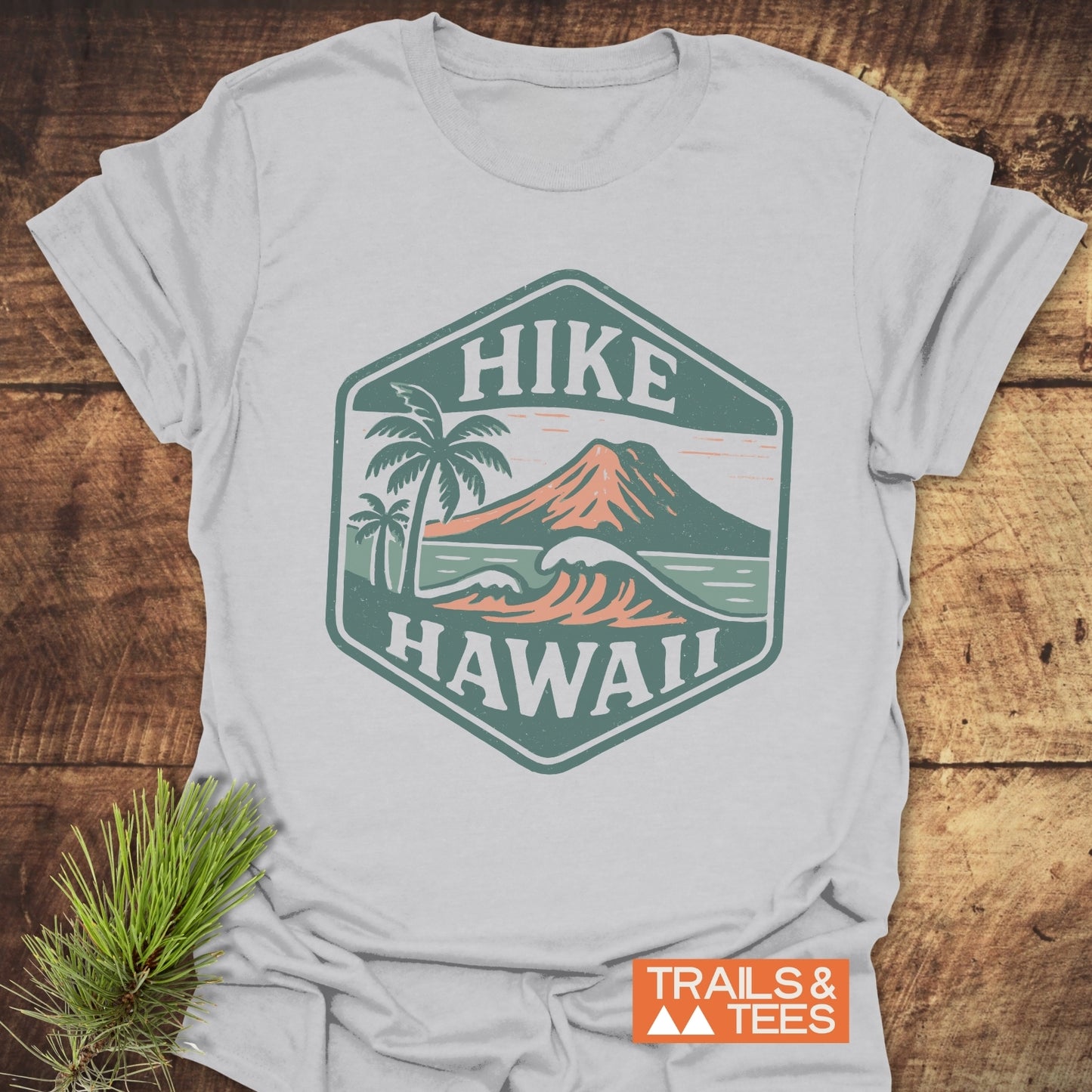 Hike Hawaii - Softstyle T-Shirt features a hexagon graphic of a volcano over ocean waves with palm trees, labeled “HIKE HAWAII,” on a light-grey tee, showcasing Trails & Tees’ adventurous spirit.