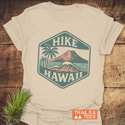 Hike Hawaii - Softstyle T-Shirt featuring a hexagonal badge with palm trees, ocean waves, and a volcanic mountain. Displayed on a wooden background with Trails & Tees logo and evergreen sprig.