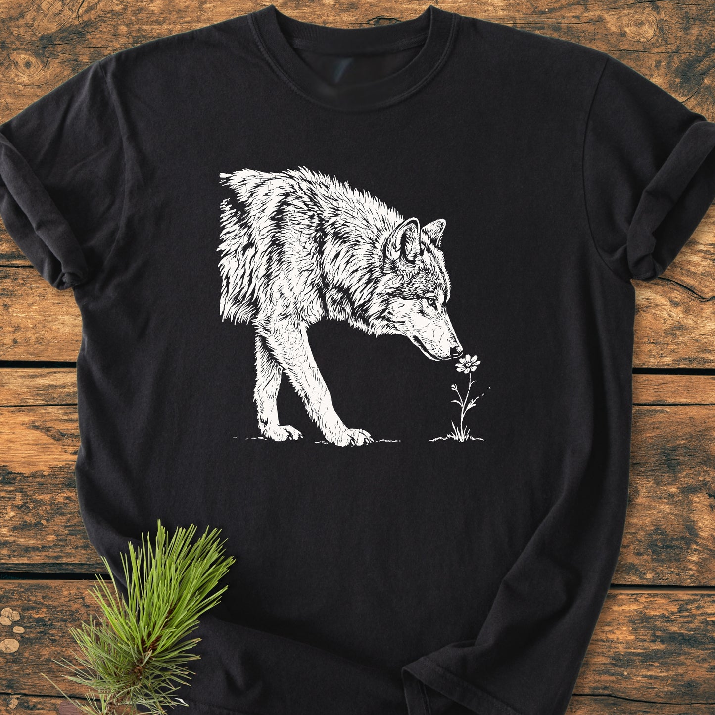 Black long-sleeve T-shirt titled Stop And Smell The Flowers - Vintage Wash Tee, featuring a white sketch of a wolf sniffing a flower, laid on rustic wood with pine needles.