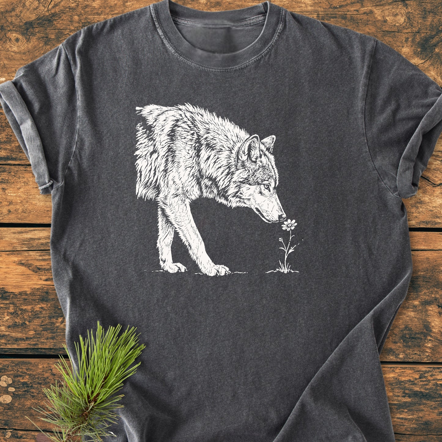 Charcoal-gray Stop And Smell The Flowers - Vintage Wash Tee featuring a white wolf sniffing a flower, laid flat on a wooden surface with pine needles for styling.