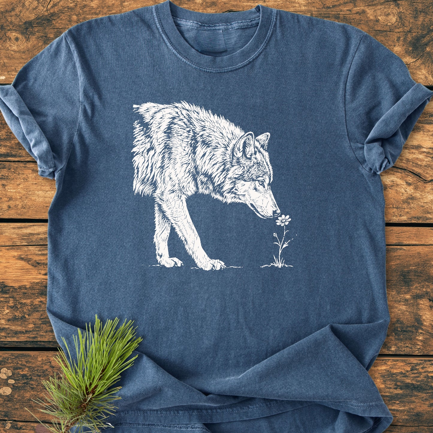 Stop And Smell The Flowers - Vintage Wash Tee featuring a wolf sniffing a flower, laid flat on wood with a pine branch accent, capturing nature's essence with a whimsical touch.