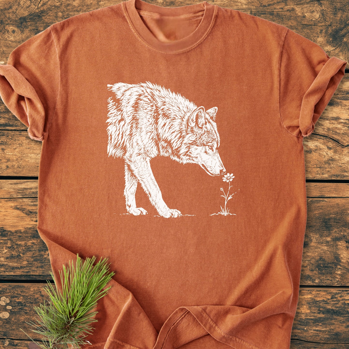 Stop And Smell The Flowers - Vintage Wash Tee featuring a white wolf sniffing a flower, styled with a pine sprig under the sleeve, laid on a wooden background.