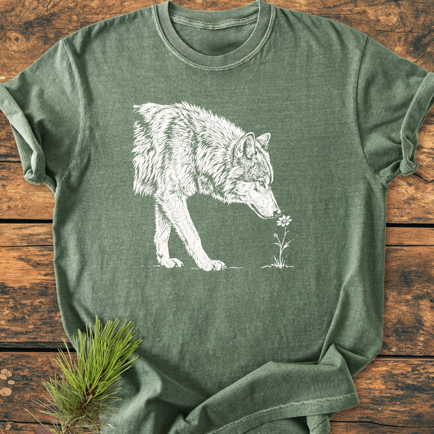 Stop And Smell The Flowers - Vintage Wash Tee displays a wolf sniffing a flower, laid flat with pine sprigs on a rustic surface, emphasizing a nature-inspired, relaxed outdoor aesthetic.