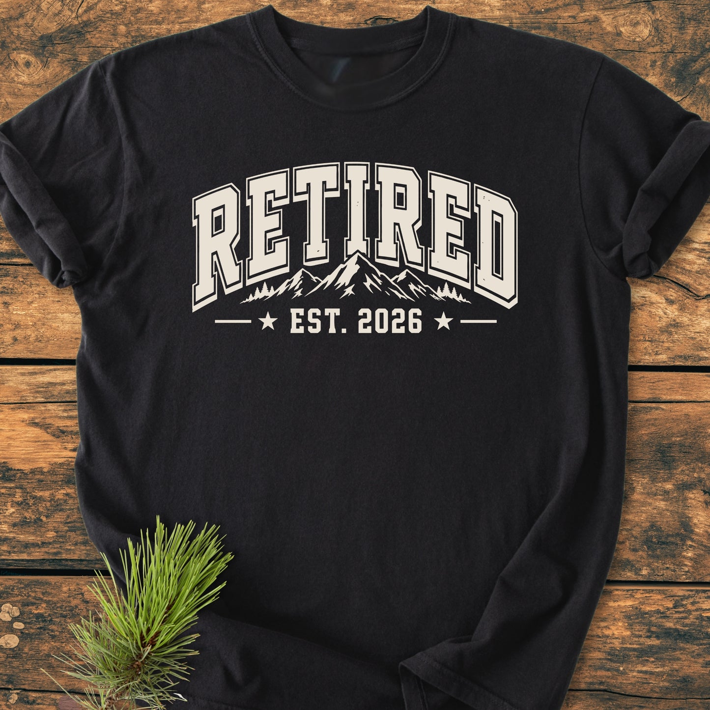 Retired 2026 - Vintage Wash Tee: Black long-sleeve shirt with “RETIRED” in collegiate print, mountain and tree graphic, “EST. 2026” below; laid on wood, pine sprig beside.
