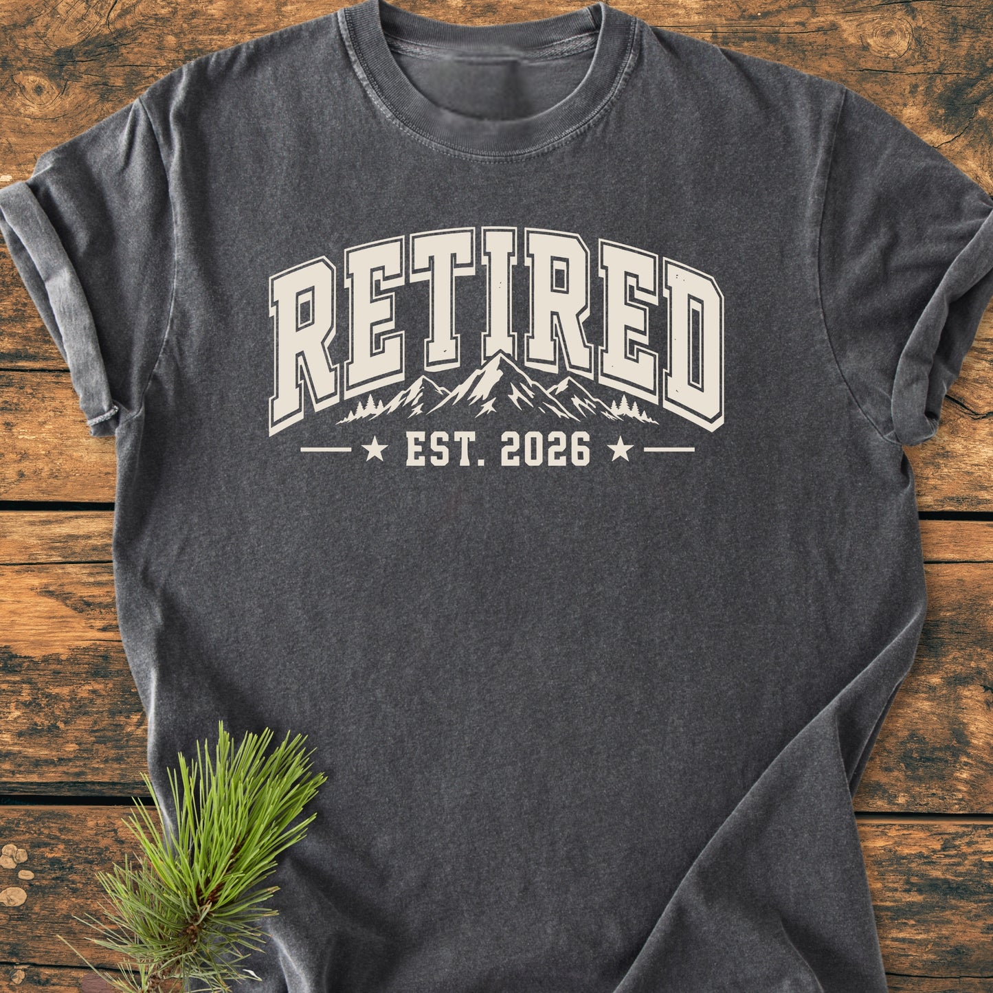 Retired 2026 - Vintage Wash Tee: Charcoal-gray T-shirt with “RETIRED” and a mountain graphic, set on rustic wood with pine sprigs, evoking an outdoorsy, seasonal vibe.