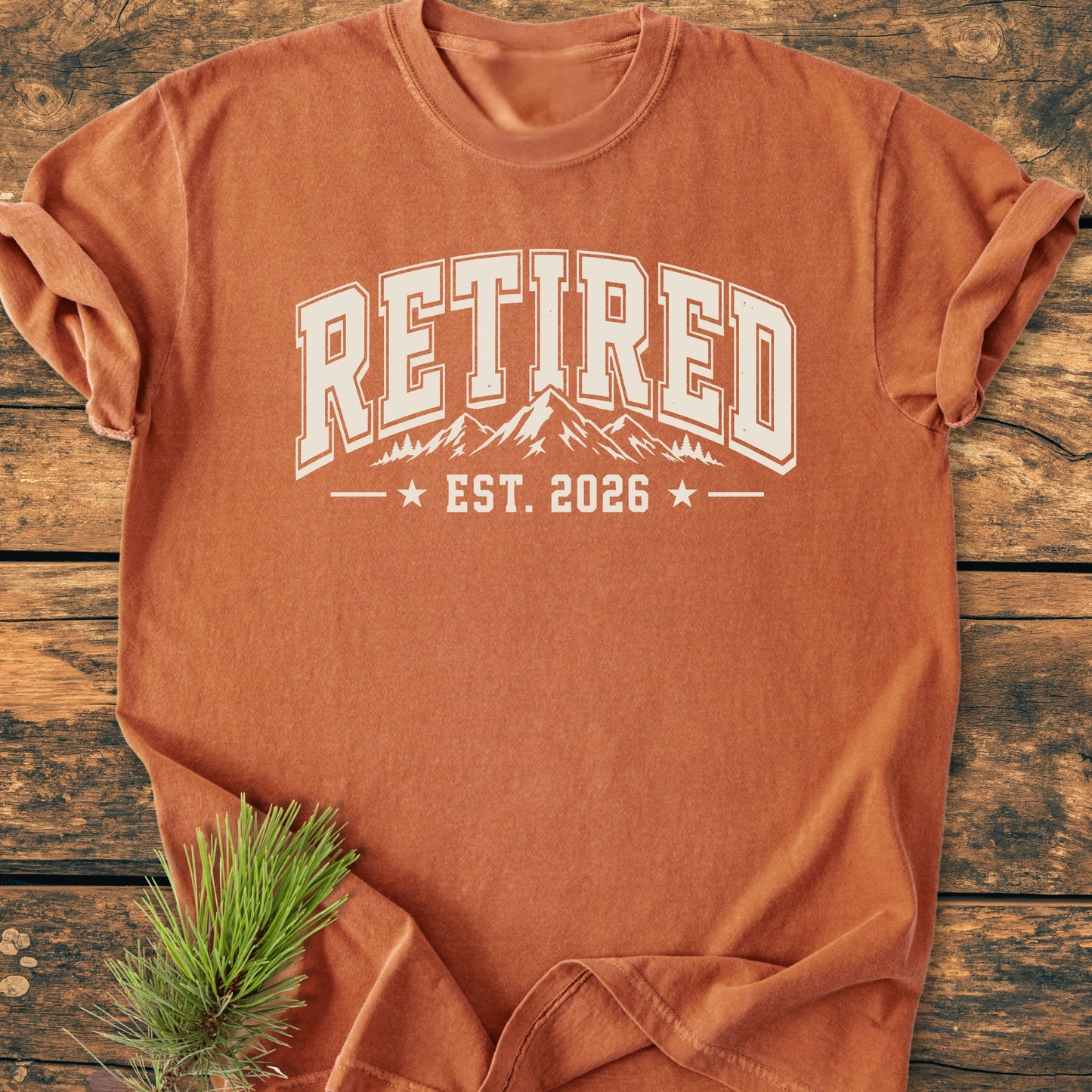 Retired 2026 - Vintage Wash Tee featuring large RETIRED text and mountain graphic, laid flat on wood; pine needles beside, embodying a relaxed, outdoor spirit perfect for post-work adventures.