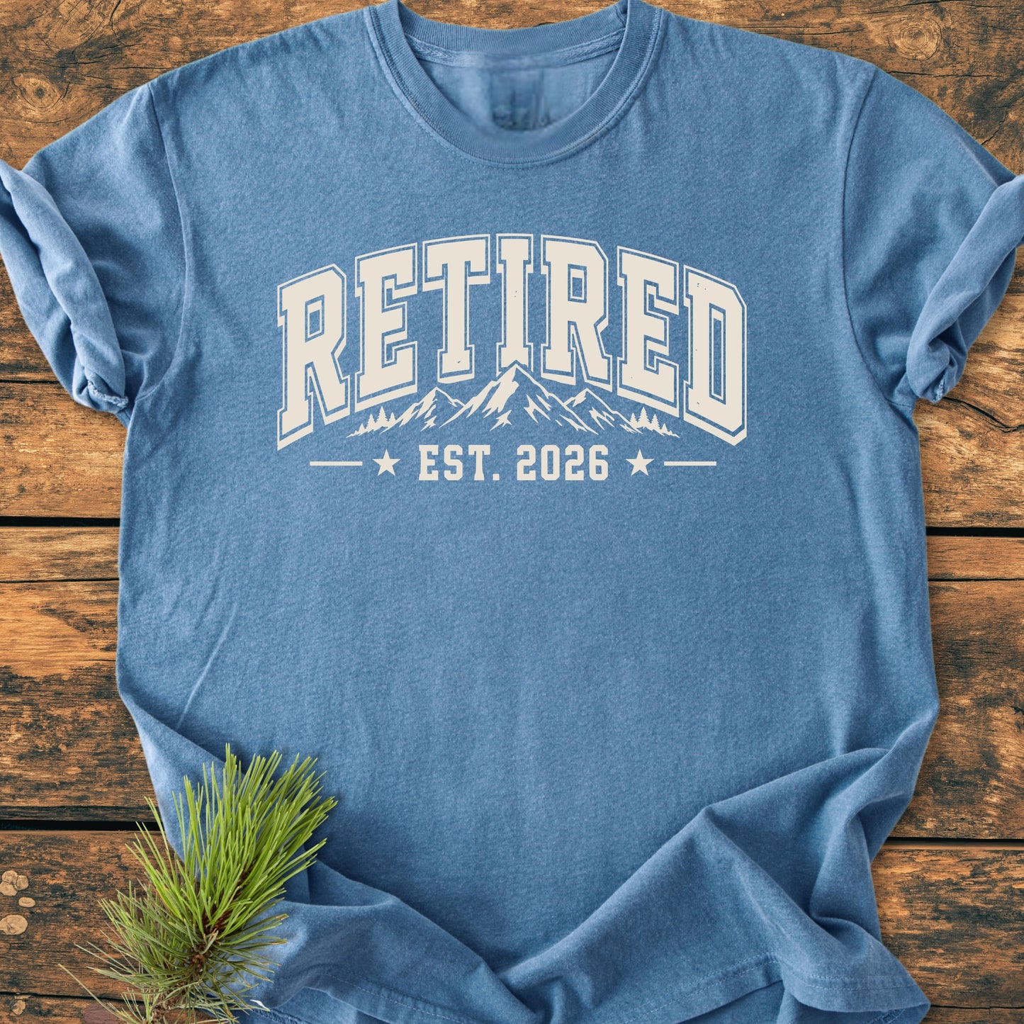 Retired 2026 - Vintage Wash Tee in dusty-blue with RETIRED collegiate graphic, mountain range, and EST. 2026. Laid on a wooden surface with pine sprig accentuating its outdoor theme.