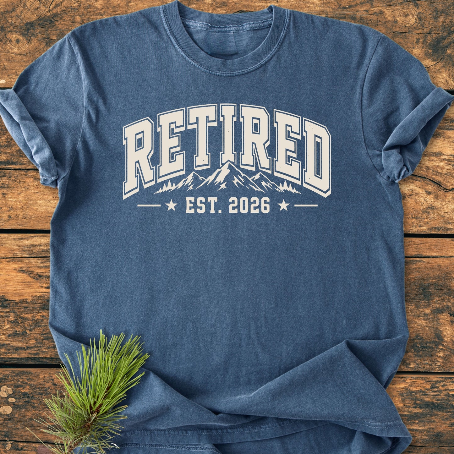 Heather-blue Retired 2026 - Vintage Wash Tee with collegiate-style RETIRED text and mountain graphic, laid flat on wood. Pine sprig accents, embodying a relaxed, outdoor-inspired vibe.