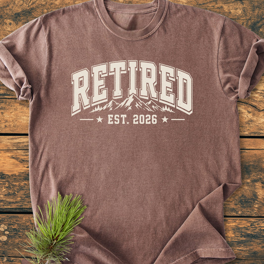 Retired 2026 - Vintage Wash Tee with collegiate print, mountain and tree line-drawing, on rustic wood. Pine bough accent. Perfect for retirees and mountain lovers embracing outdoor adventures.