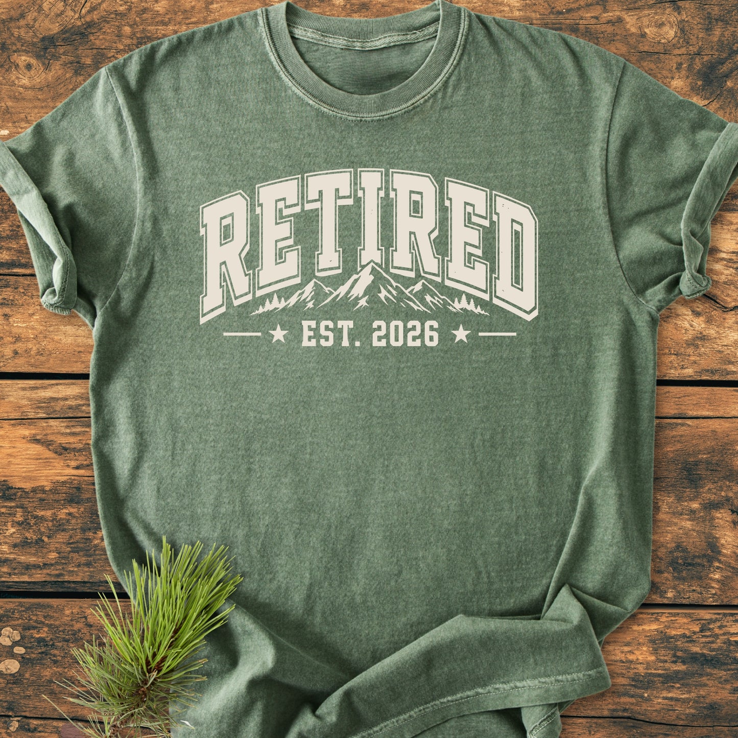 Heather-green Retired 2026 - Vintage Wash Tee with cream collegiate graphic and mountain silhouette, laid on wood, evokes an outdoorsy, retirement vibe. Accompanied by a pine needle sprig.