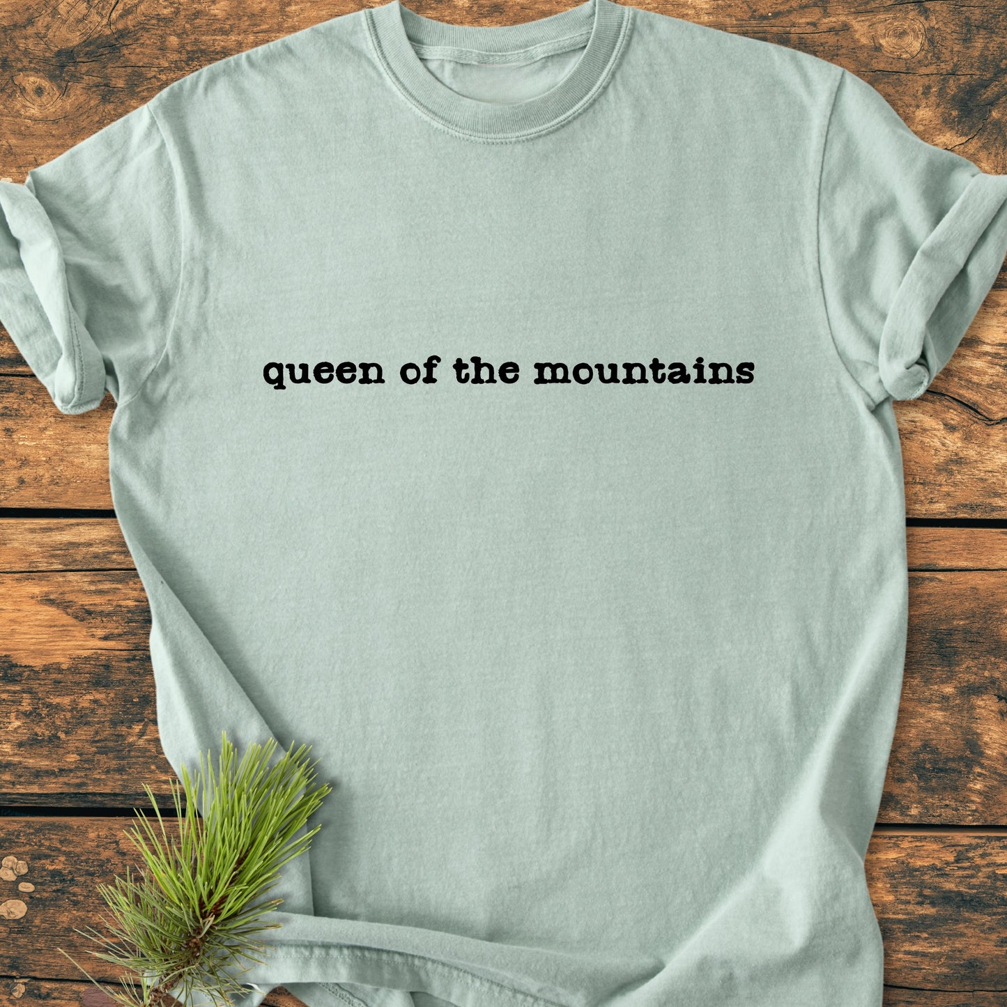 Queen Of The Mountains - Vintage Wash Tee displayed on a wooden surface, featuring typewriter-style text across the chest. Enhanced with a pine sprig for a rustic feel, embodying mountain-inspired elegance.