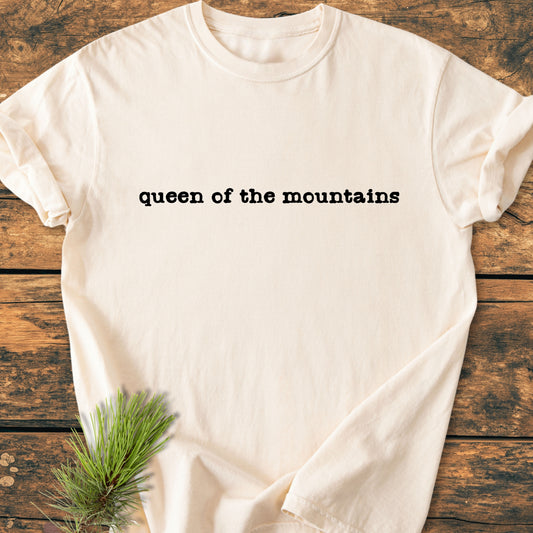 Queen Of The Mountains - Vintage Wash Tee, cream-colored with black typewriter font, displayed flat on wood. A sprig of pine adds an outdoorsy touch, emphasizing its rustic, natural appeal.