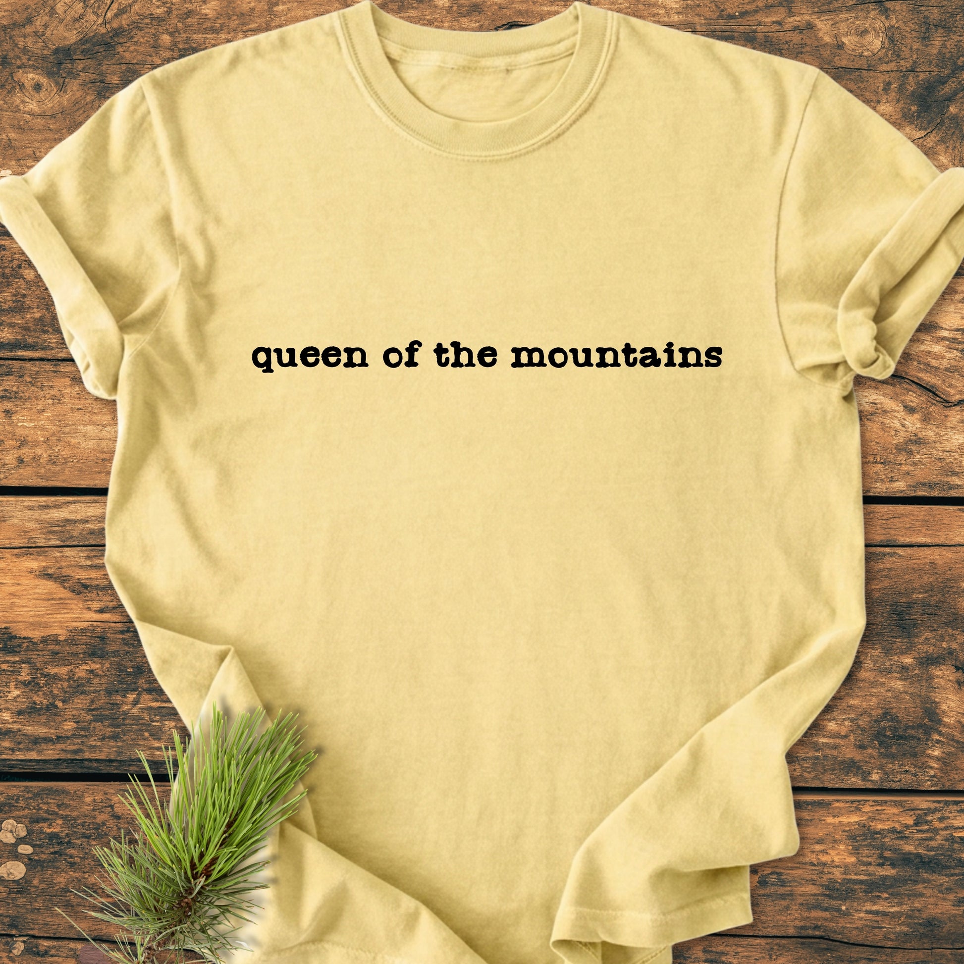 Pale yellow Queen of the Mountains T-shirt in black typewriter-style font, flat-laid on rustic wood, accented by a pine sprig, embodying a nature-inspired, relaxed fit vintage wash tee.