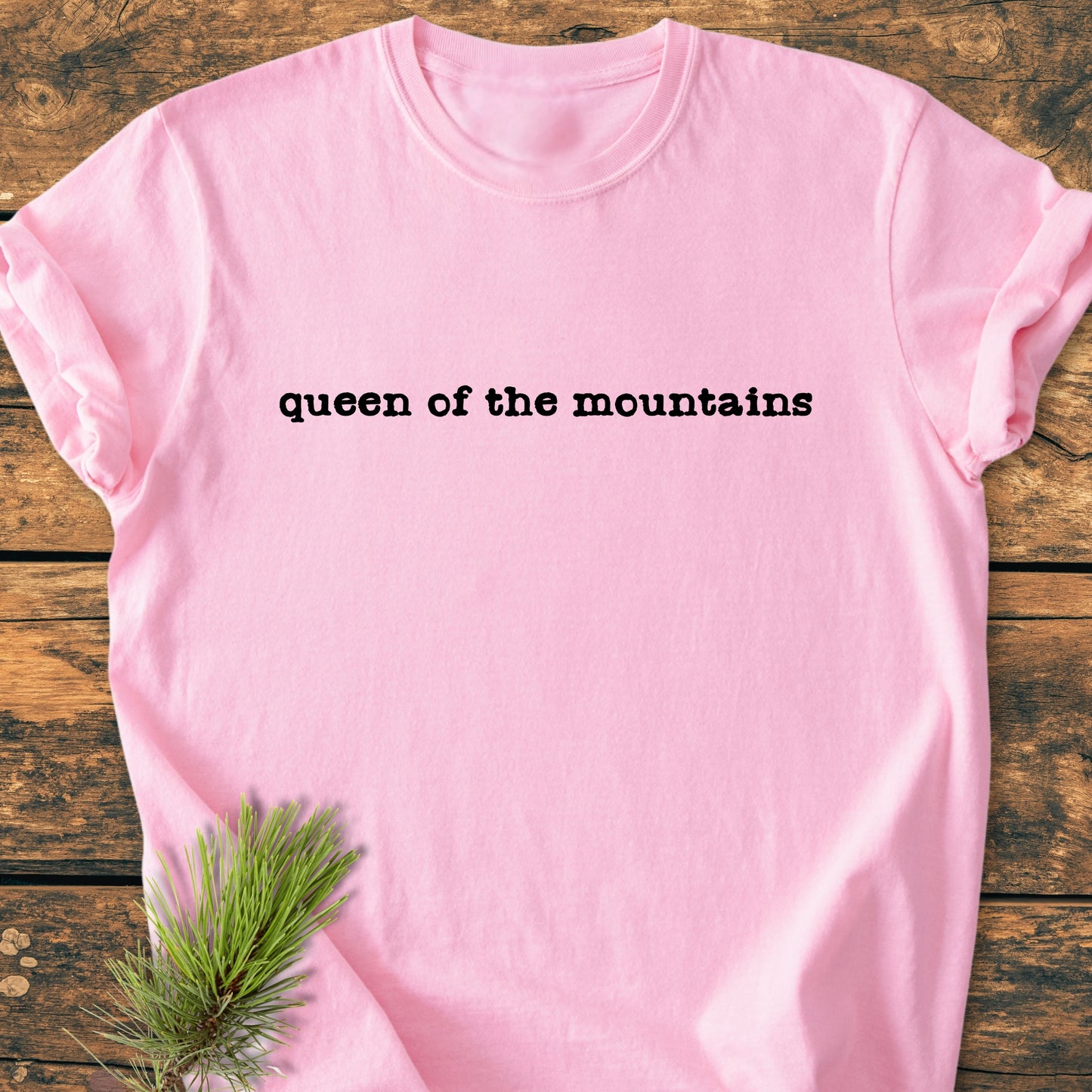 Queen Of The Mountains - Vintage Wash Tee laid flat on rustic wood, featuring black typewriter-style text. A pine branch sprig accents the scene, highlighting the tee's outdoor mountain-inspired aesthetic.