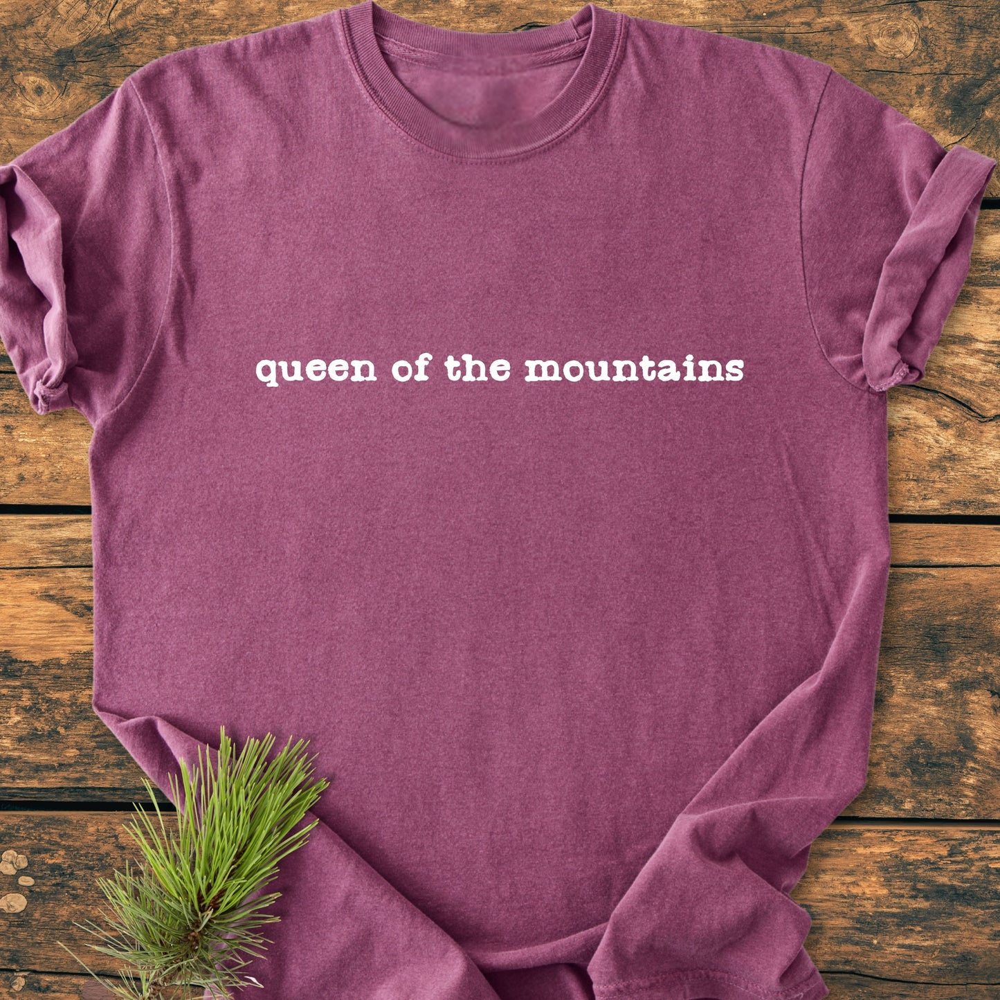 Queen Of The Mountains - Vintage Wash Tee: Mauve-purple crew-neck T-shirt on wooden surface, featuring “queen of the mountains” slogan in white font, with pine sprig accentuating its outdoor theme.