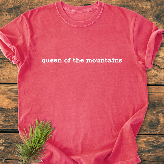 Queen Of The Mountains - Vintage Wash Tee, a red crew-neck T-shirt with white stenciled text, laid on a wooden surface with a pine sprig beside it, symbolizing outdoor strength and tranquility.