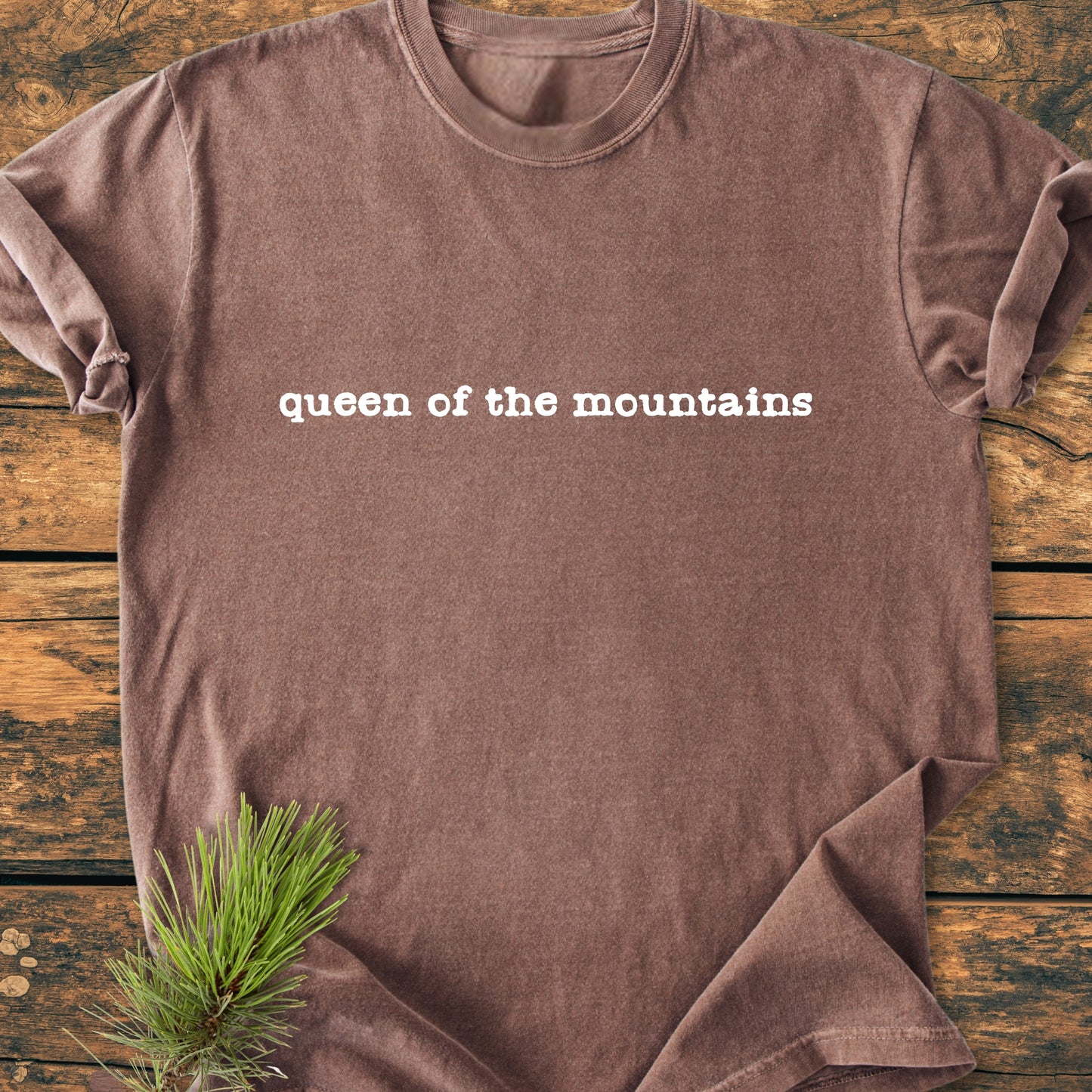 Queen Of The Mountains - Vintage Wash Tee laid on rustic wood, features white typewriter-style text, and is accented with a green pine branch, highlighting its outdoor-inspired design.