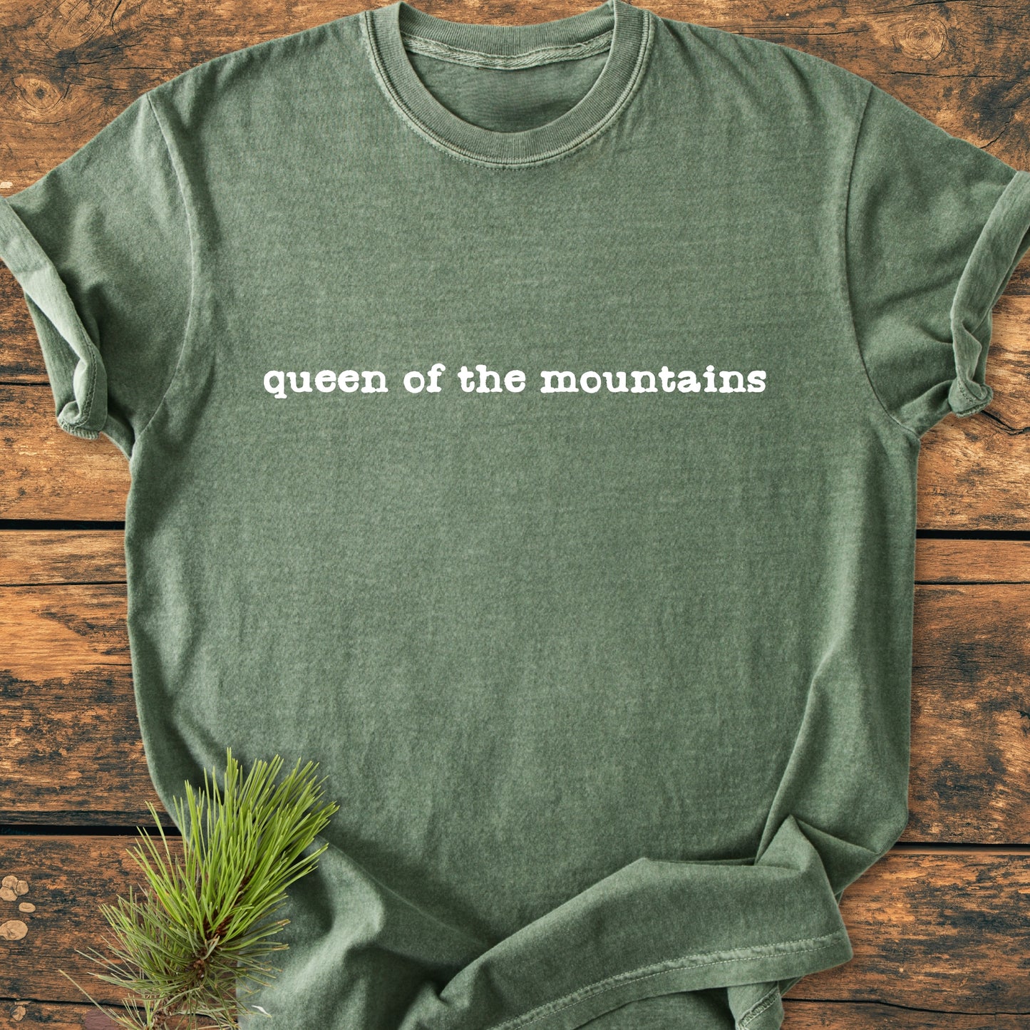 Heathered green Queen Of The Mountains - Vintage Wash Tee laid on wooden surface, featuring white typewriter-style text. A pine branch peeks from under the hem, emphasizing its outdoorsy theme.