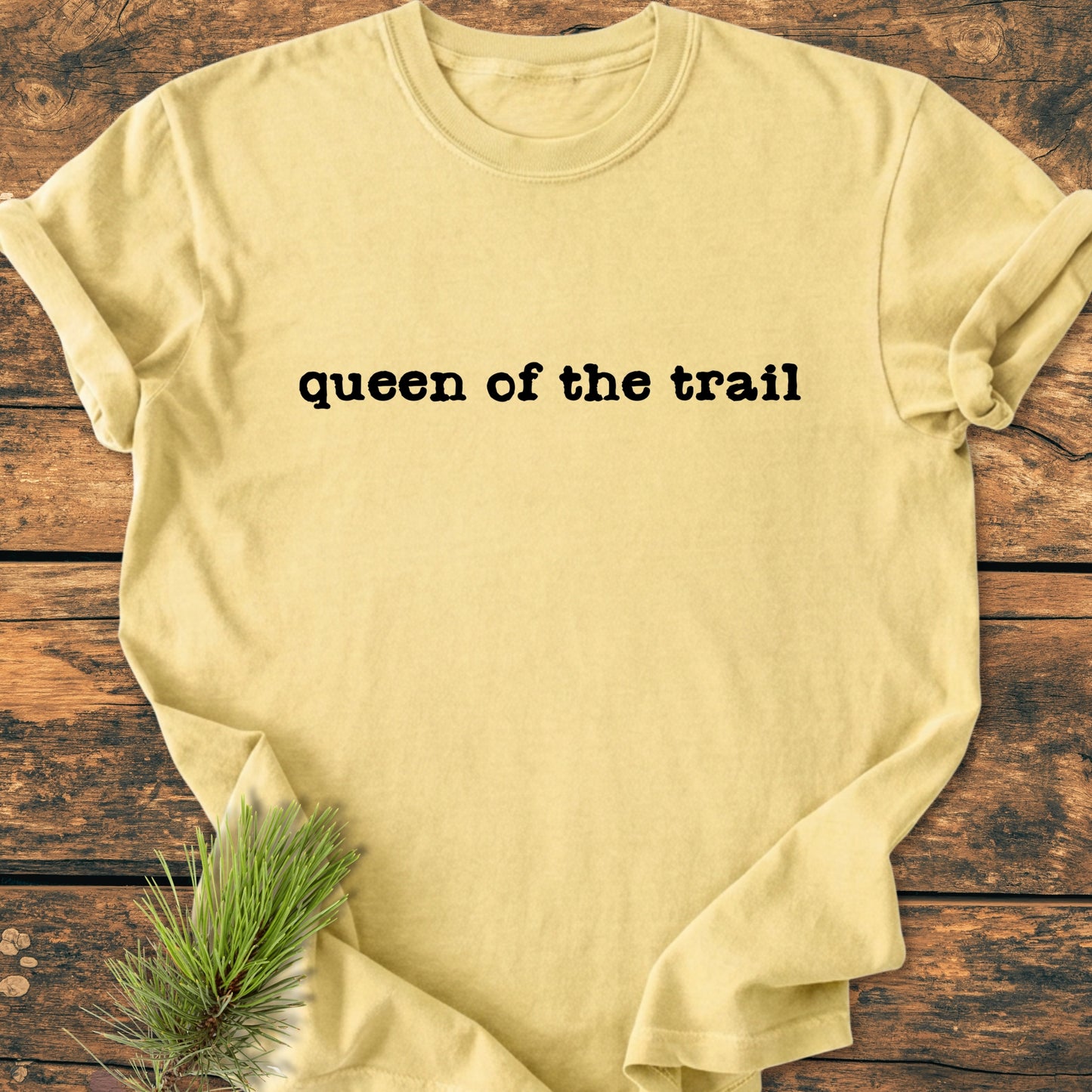 Queen Of The Trail - Vintage Wash Tee displayed on wood, featuring bold black lettering and pine needles, highlighting its trail-inspired theme and relaxed, outdoor-loving vibe.