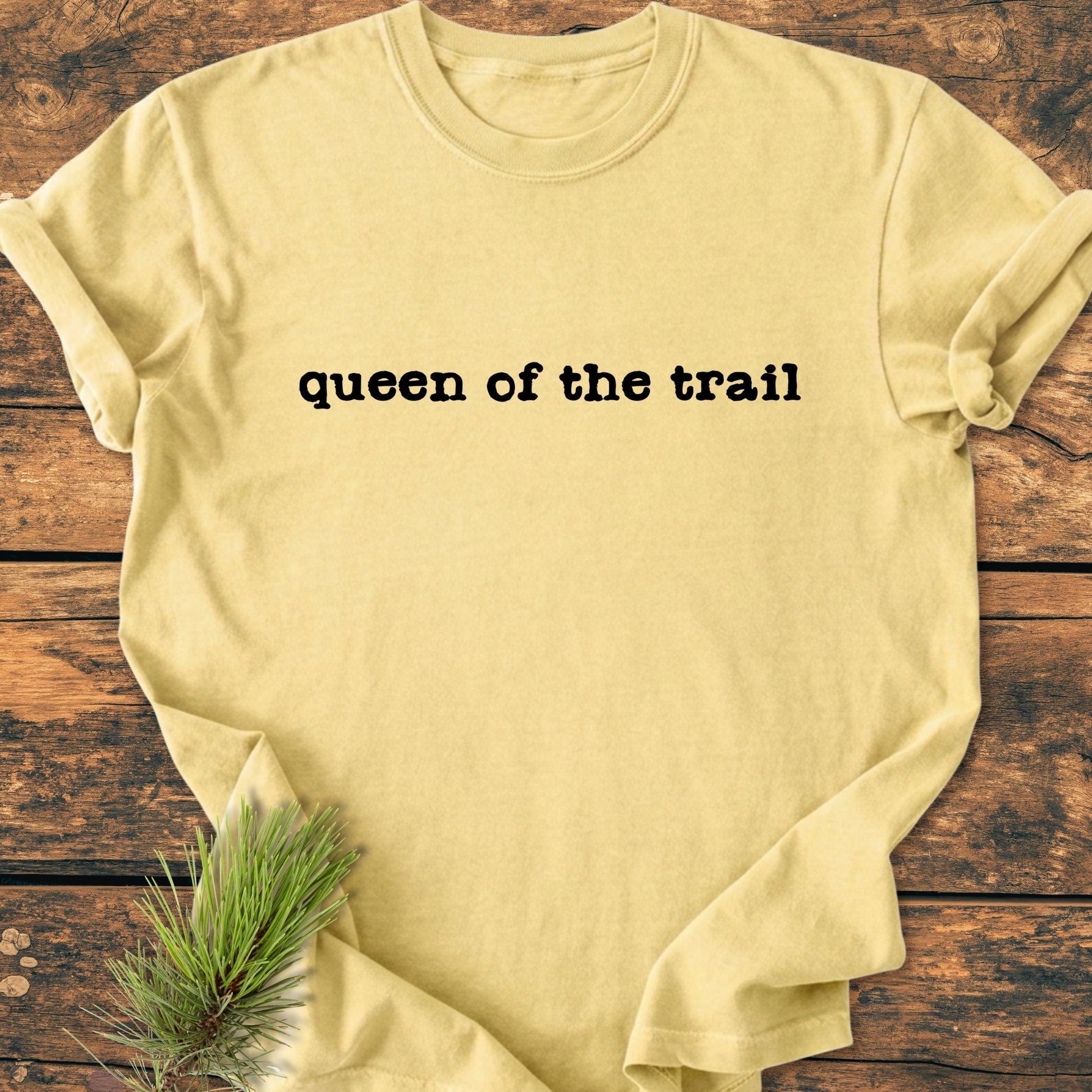 Queen Of The Trail - Vintage Wash Tee displayed on wood, featuring bold black lettering and pine needles, highlighting its trail-inspired theme and relaxed, outdoor-loving vibe.