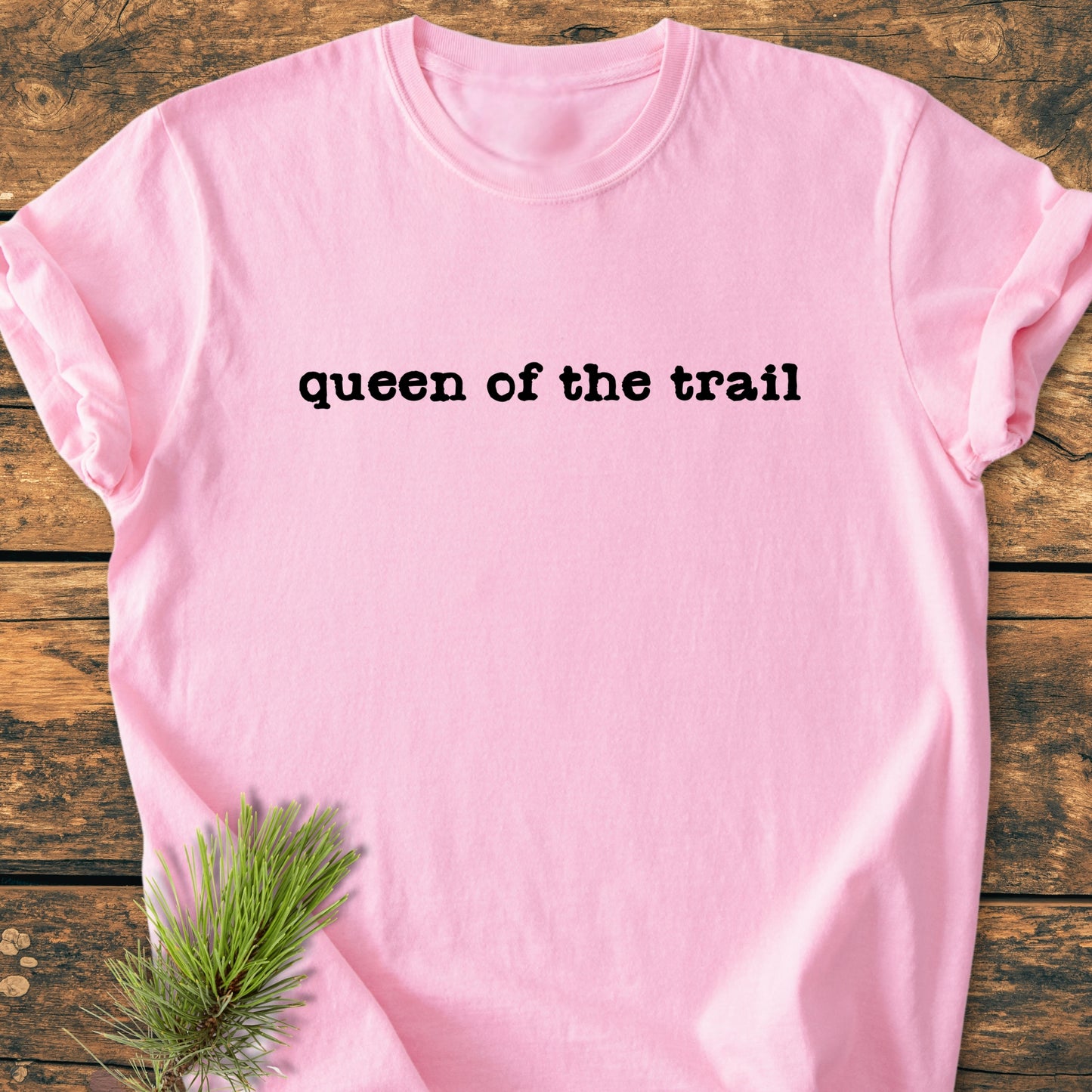 Light-pink tee with queen of the trail in black typewriter font, laid flat on wooden surface. Pine needles accent the corner, emphasizing the vintage wash's outdoor inspiration and relaxed fit.