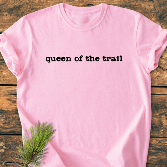Light-pink tee with queen of the trail in black typewriter font, laid flat on wooden surface. Pine needles accent the corner, emphasizing the vintage wash's outdoor inspiration and relaxed fit.