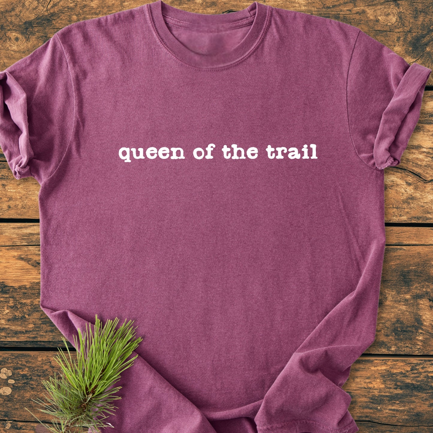 Queen Of The Trail - Vintage Wash Tee in heathered maroon, laid flat with bold white text across the chest, complemented by a pine needle sprig on a rustic wooden surface.