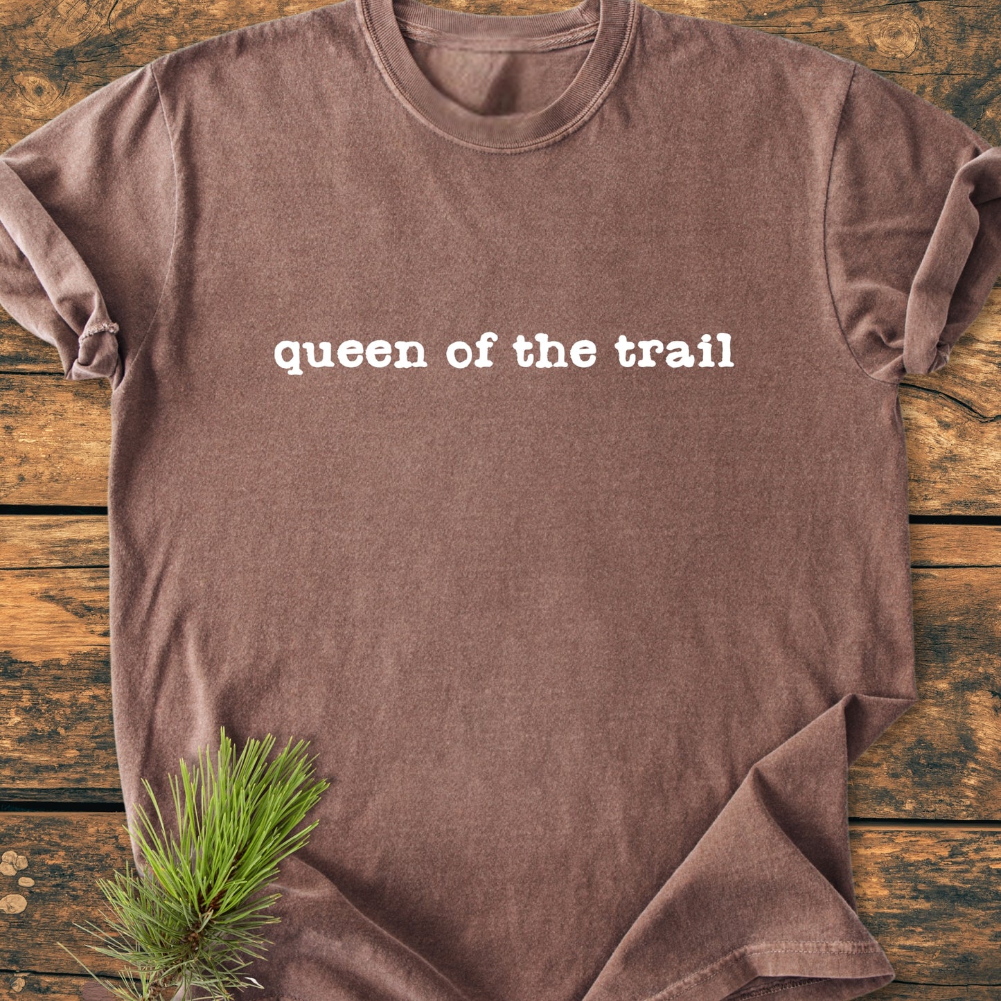 Queen Of The Trail - Vintage Wash Tee, brown short-sleeve with rolled hems and queen of the trail in white text, laid flat on a rustic wooden surface with pine sprigs.