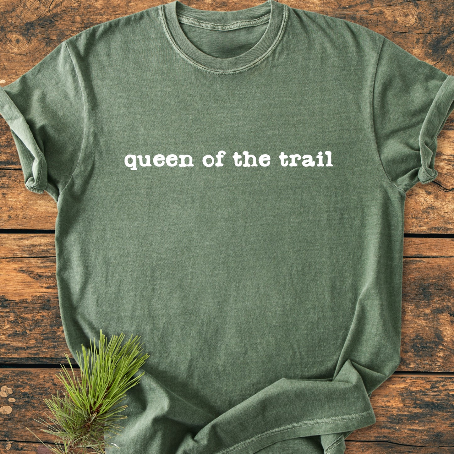 Heather-green T-shirt labeled Queen Of The Trail in white, laid on wood with a pine sprig, capturing an outdoorsy feel. Ideal for hikers, featuring a vintage wash and relaxed fit.
