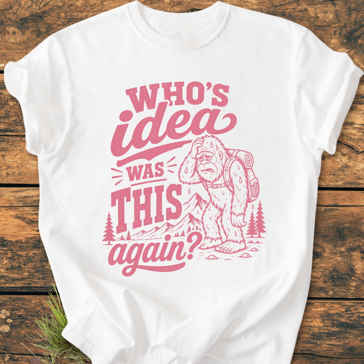 White T-shirt featuring Who's Idea Bigfoot - Vintage Wash Tee with a cartoon Bigfoot carrying a backpack, surrounded by trees and mountains, and the phrase WHO’S IDEA WAS THIS again? in vintage lettering.