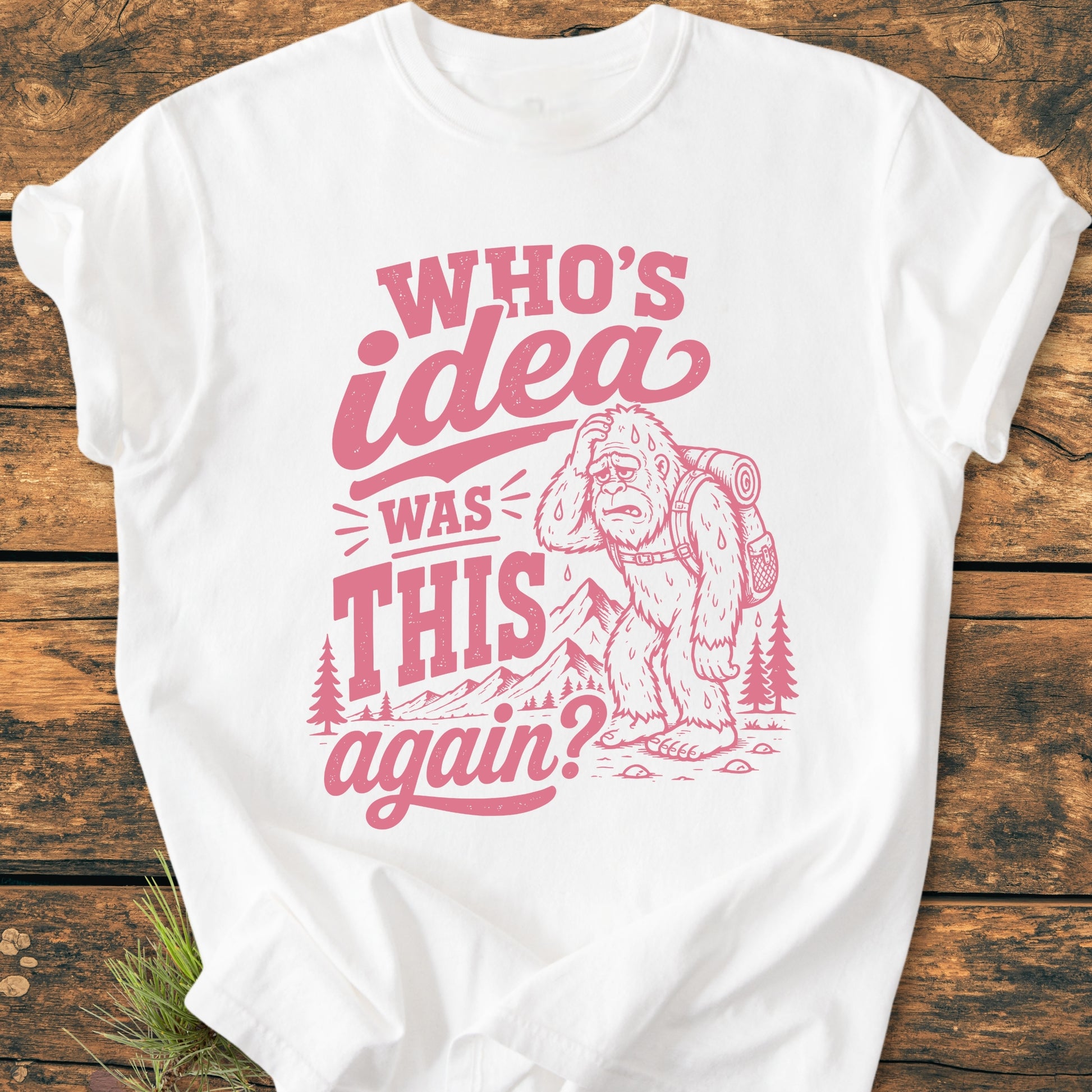 White T-shirt featuring Who's Idea Bigfoot - Vintage Wash Tee with a cartoon Bigfoot carrying a backpack, surrounded by trees and mountains, and the phrase WHO’S IDEA WAS THIS again? in vintage lettering.