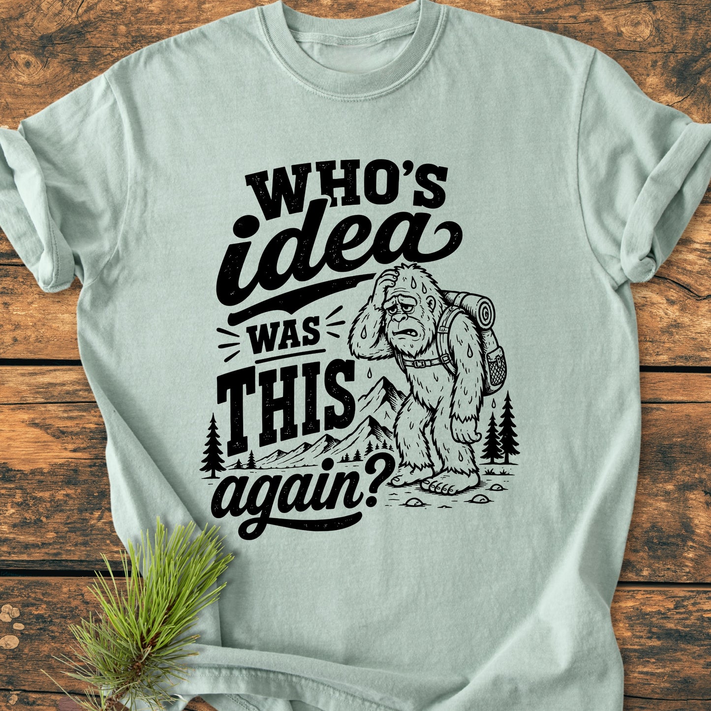 Pale-green tee featuring Who's Idea Bigfoot - Vintage Wash Tee with a cartoon Bigfoot hiking design and humorous text, laid on a wooden plank, accompanied by a pine bough.