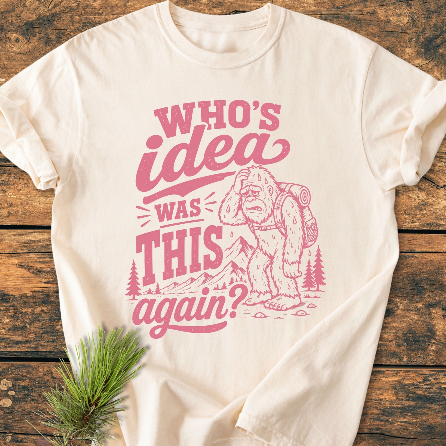 Light-cream T-shirt titled Who's Idea Bigfoot - Vintage Wash Tee features a pink vintage graphic with a cartoon Bigfoot, mountains, and trees, symbolizing humorous hiking mishaps.