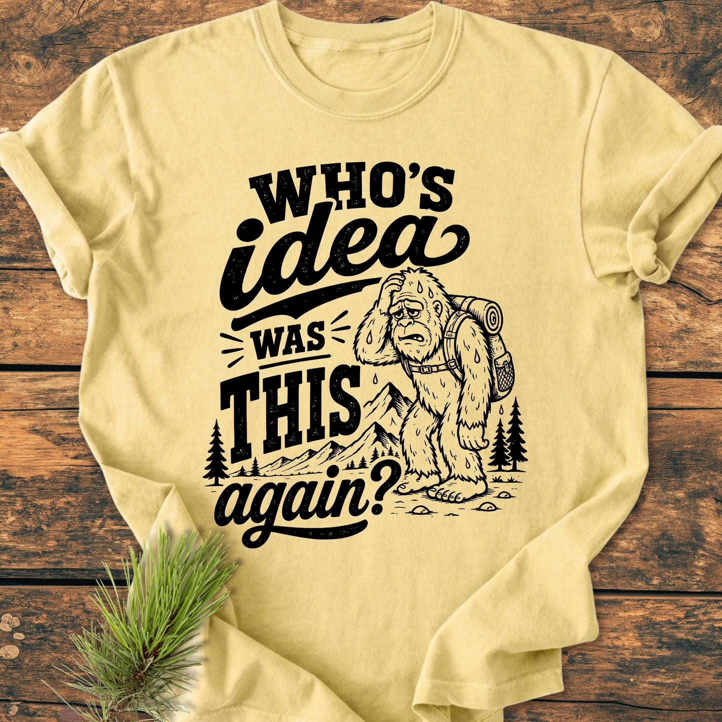 Who's Idea Bigfoot - Vintage Wash Tee features a comic Bigfoot, backpacking and puzzled, on a pale-yellow long-sleeve, capturing the humor of outdoor adventures with a mountain backdrop.