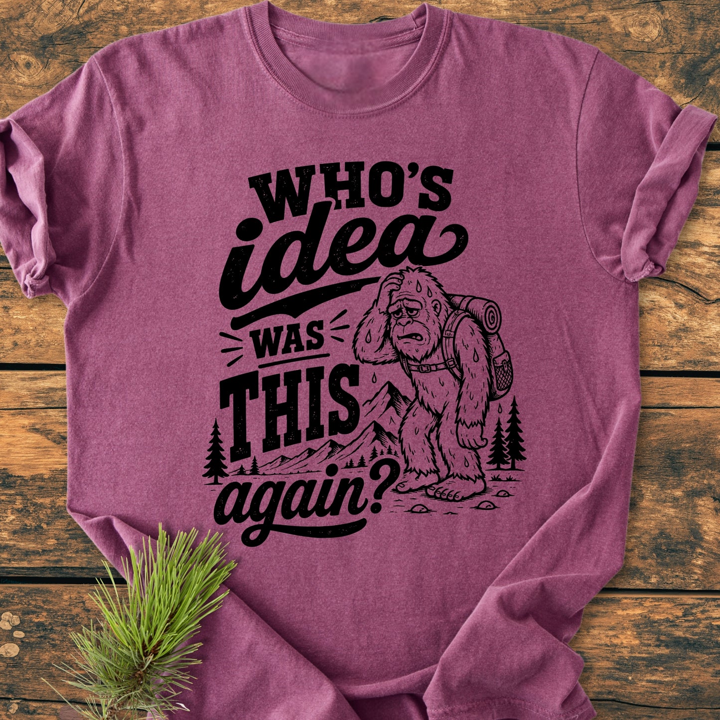 The Who's Idea Bigfoot - Vintage Wash Tee features a cartoon Bigfoot hiking with text, laid on rustic wood, capturing the humor and adventure spirit for outdoor enthusiasts.
