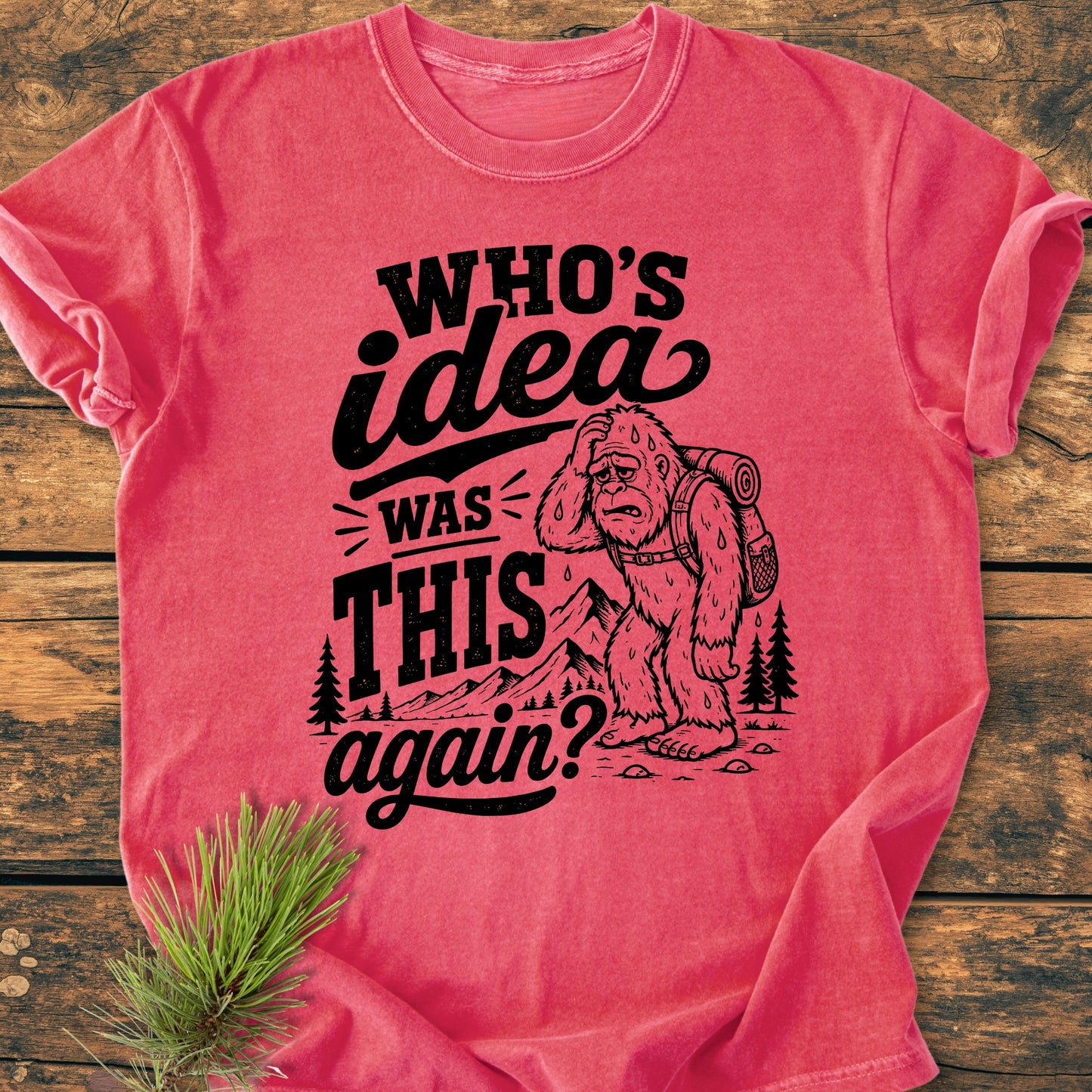 Red T-shirt featuring a black graphic of a tired Bigfoot with a backpack, surrounded by mountains and trees. Bold text reads, “WHO’S idea was THIS again?” Perfect for hikers and adventure lovers.