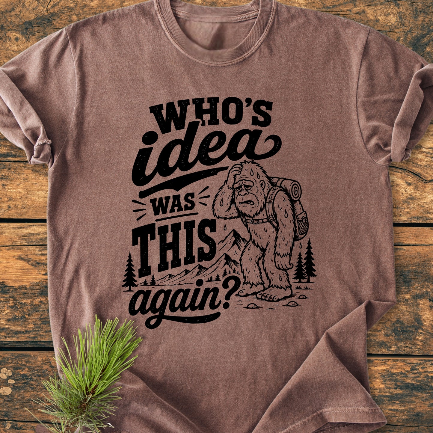 Who's Idea Bigfoot - Vintage Wash Tee featuring a cartoon Bigfoot in front of a mountain, laid on wooden surface with pine sprig, capturing the humor and exhaustion of hiking adventures.