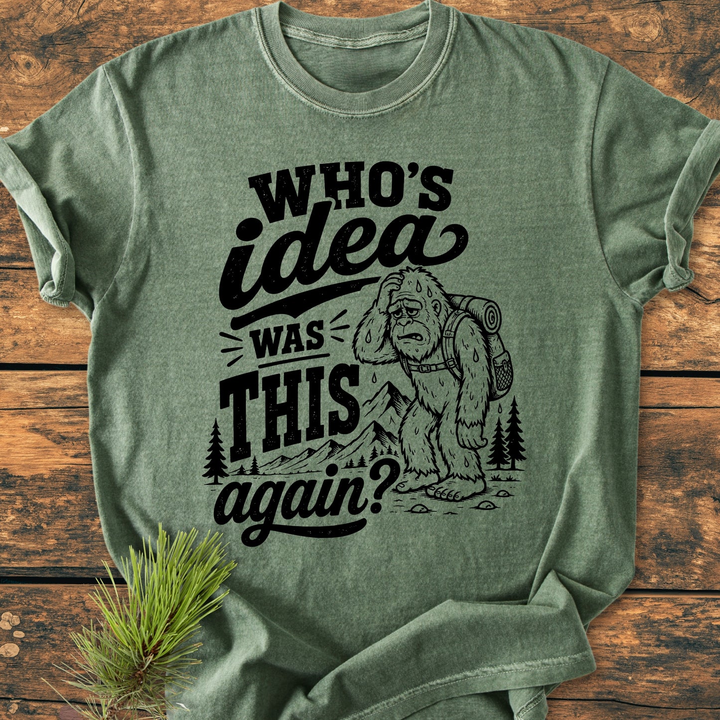 Who's Idea Bigfoot - Vintage Wash Tee features a cartoon sasquatch, exhausted with a backpack, stumbling past trees and mountains on a soft, garment-dyed, heathered green T-shirt.