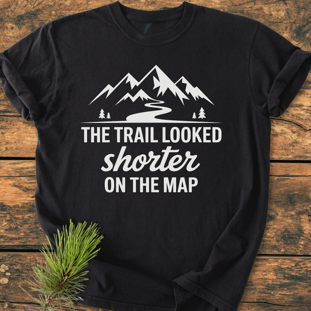 Black T-shirt with mountain peaks, trail, pine trees graphic, and The Trail Looked Shorter On The Map slogan, laid on rustic wood, capturing outdoor humor in a vintage wash style.