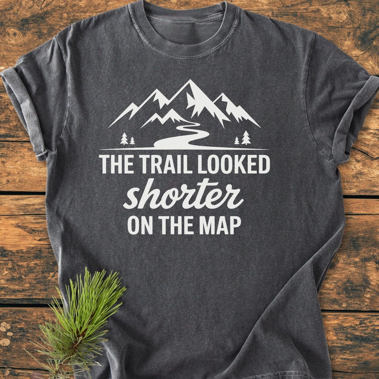 Charcoal-gray T-shirt with white graphic mountains and trail, captioned “THE TRAIL LOOKED shorter ON THE MAP.” Features pine trees, suited for hikers seeking humor in underestimating trails.
