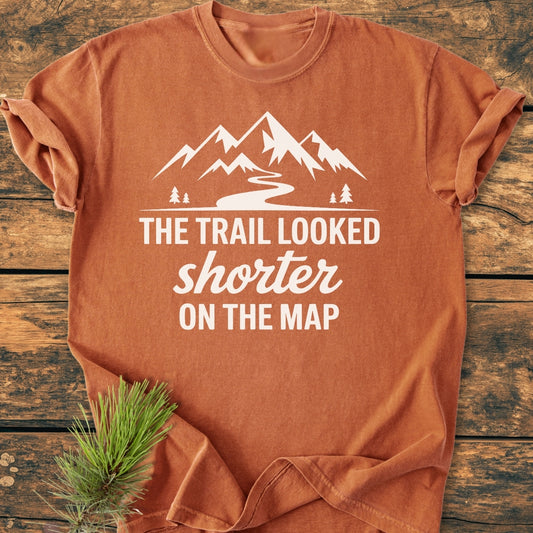 Rust-orange crew-neck T-shirt with a stylized white mountain range and trail design, captioned “The Trail Looked Shorter on the Map,” laid out on a wooden surface with a pine sprig beside it.