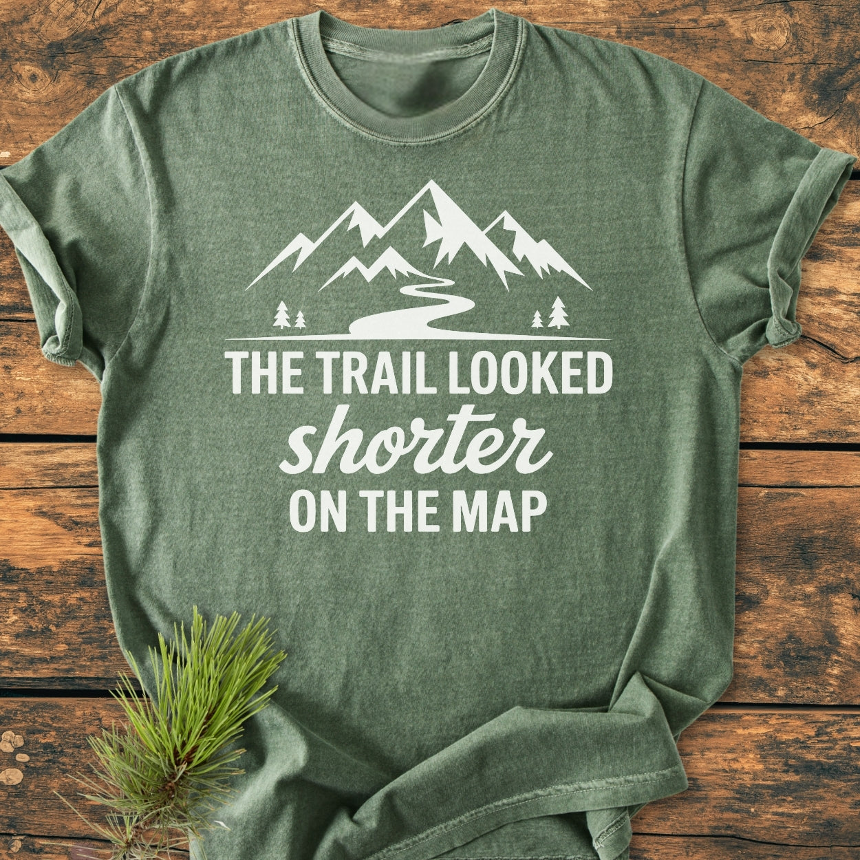 Forest-green heather T-shirt with a mountain silhouette and the slogan The Trail Looked Shorter on the Map printed in white, styled with a pine sprig on a wooden surface.