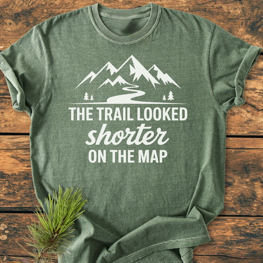 Forest-green heather T-shirt with a mountain silhouette and the slogan The Trail Looked Shorter on the Map printed in white, styled with a pine sprig on a wooden surface.