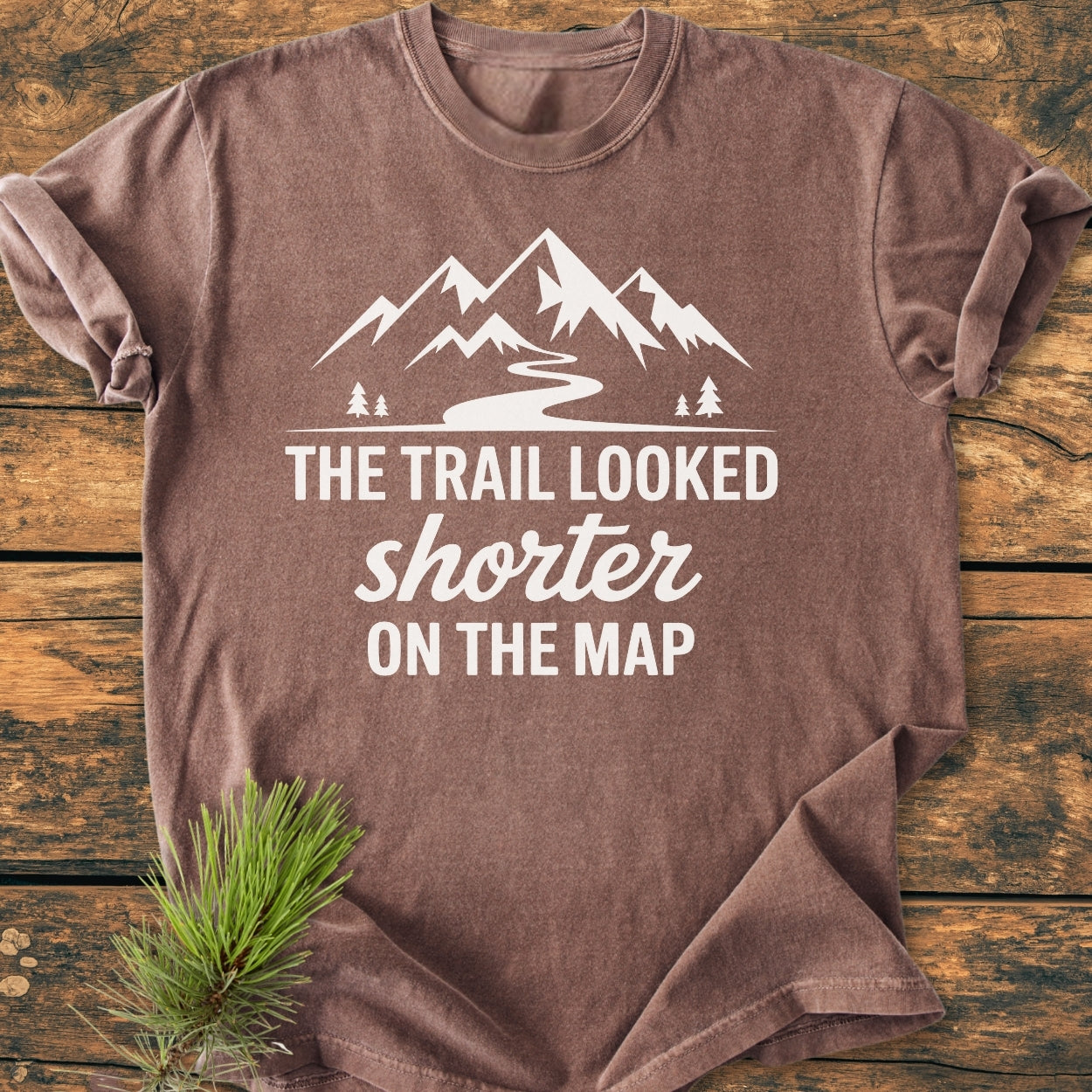 Brown short-sleeved T-shirt on wood, featuring a mountain silhouette and The Trail Looked Shorter On The Map phrase in white. Perfect for hikers appreciating trail humor and vintage wash comfort.