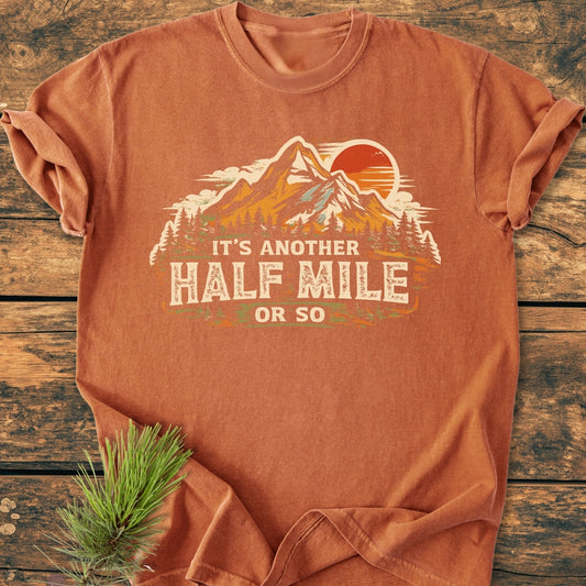 Another Half Mile Or So - Vintage Wash Tee features a stylized outdoors graphic with trees, a mountain, and sun, laid on wood surface, perfect for trail enthusiasts, offering a soft, lived-in feel.