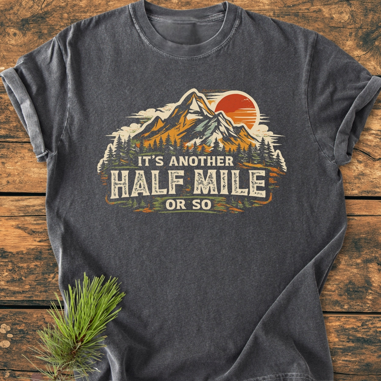 Charcoal-gray T-shirt with a mountain and forest scene, slogan It’s another HALF MILE or so, designed for hikers, laid on rustic wood. Vintage wash, relaxed fit for everyday wear.