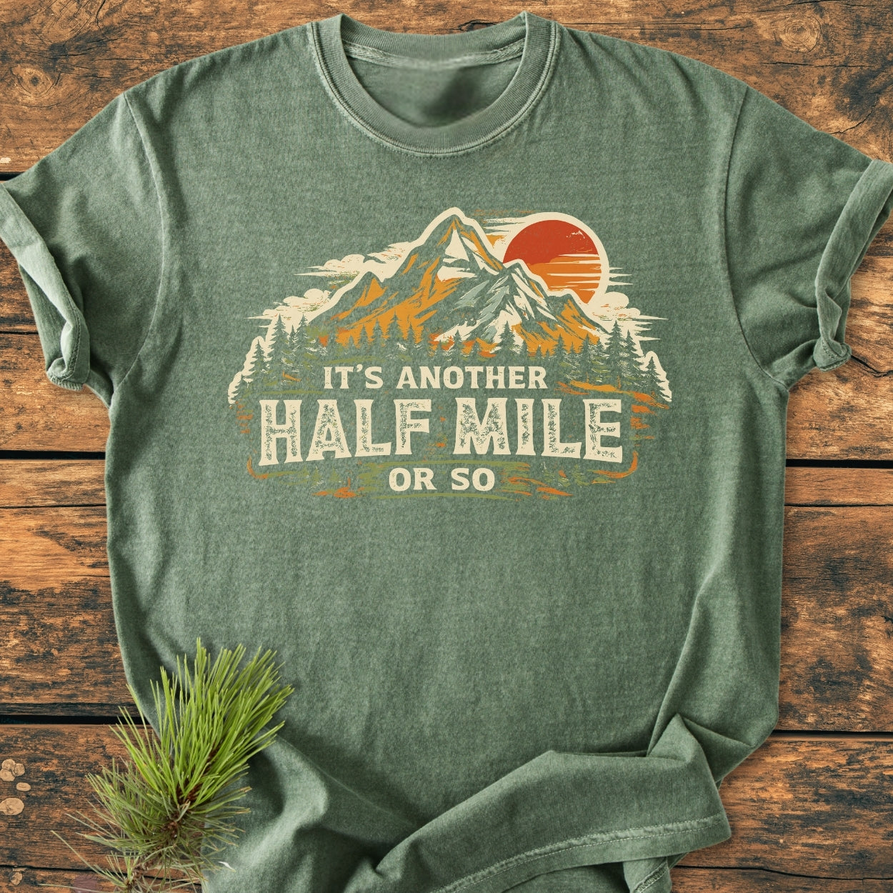 Another Half Mile Or So - Vintage Wash Tee with a mountain and pine-tree scene, red sun, and slogan; laid on wooden surface, embodies a relaxed, lived-in feel for trail enthusiasts.