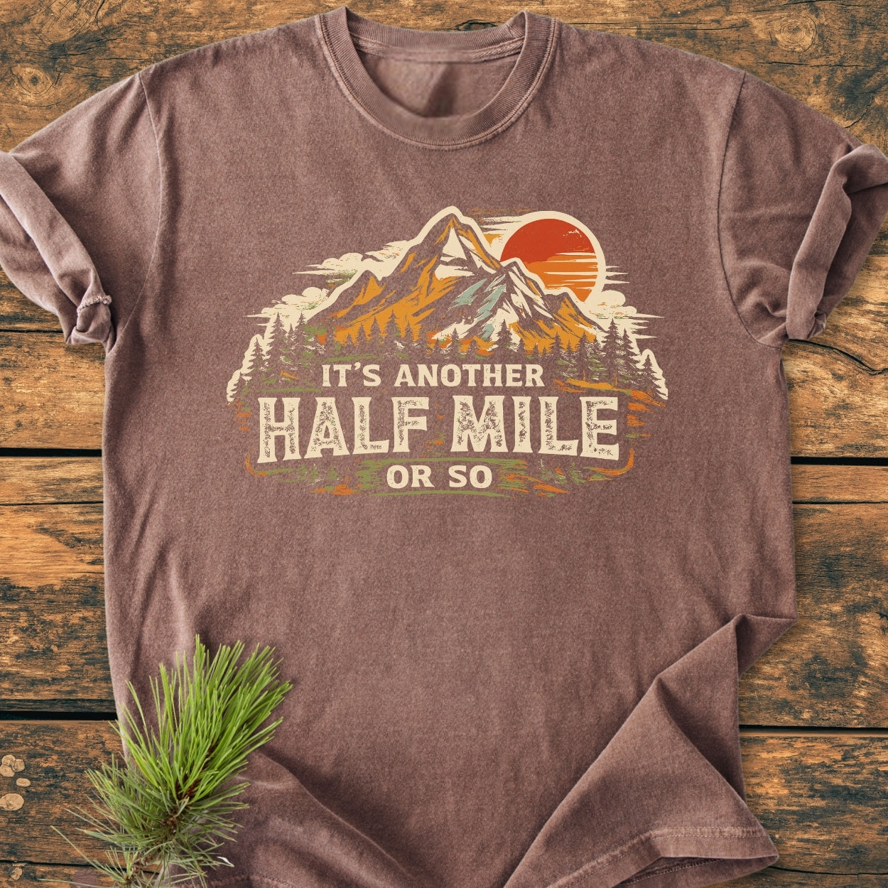 Another Half Mile Or So - Vintage Wash Tee featuring a mountain scene with peaks and pine trees, laid flat on wood, showcasing its relaxed fit and soft, lived-in feel.