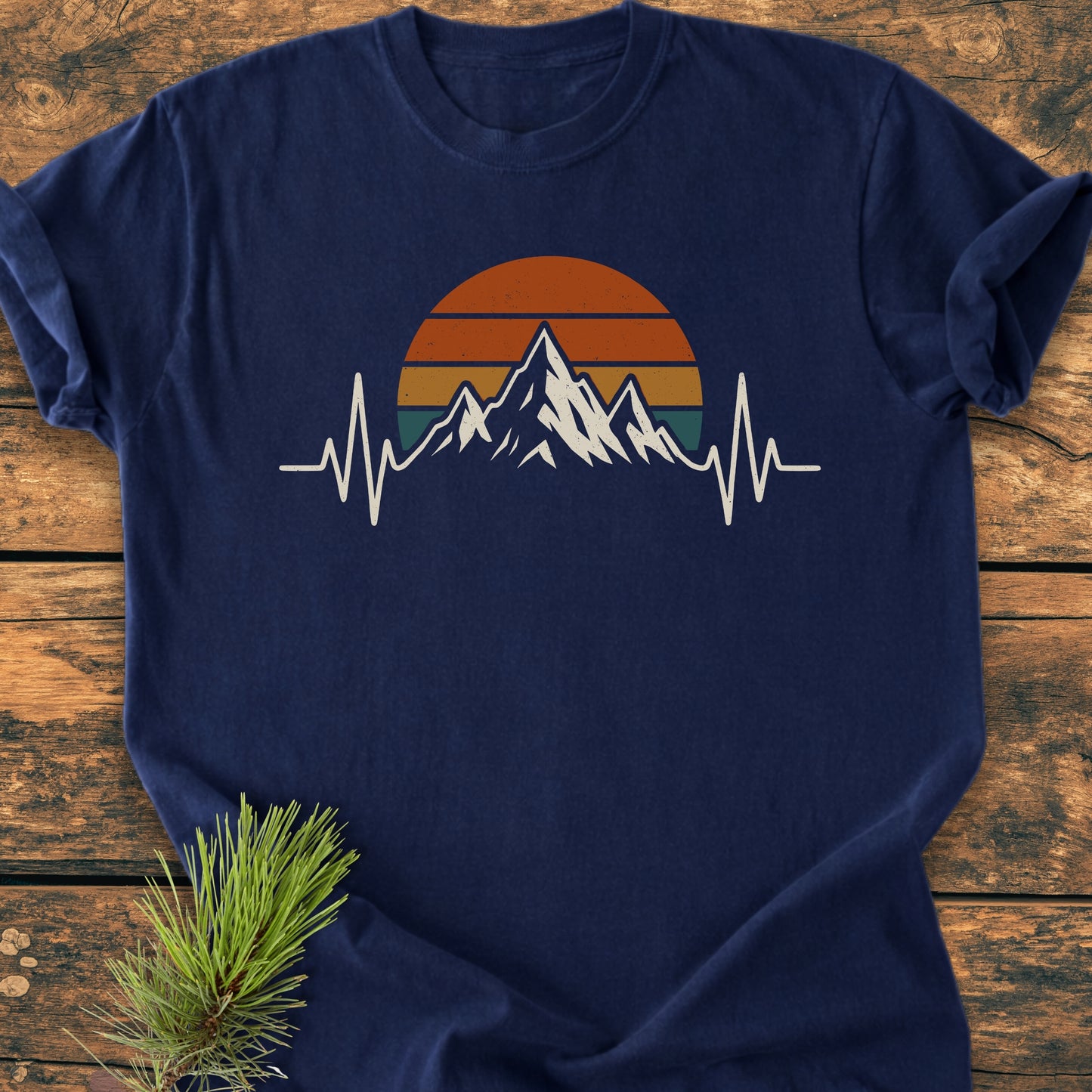 Mountain Heartbeat - Vintage Wash Tee: Navy-blue T-shirt with a stylized mountain range, half-sun, and EKG heartbeat design. Laid on wood with a pine sprig, highlighting its outdoor, trail-inspired essence.